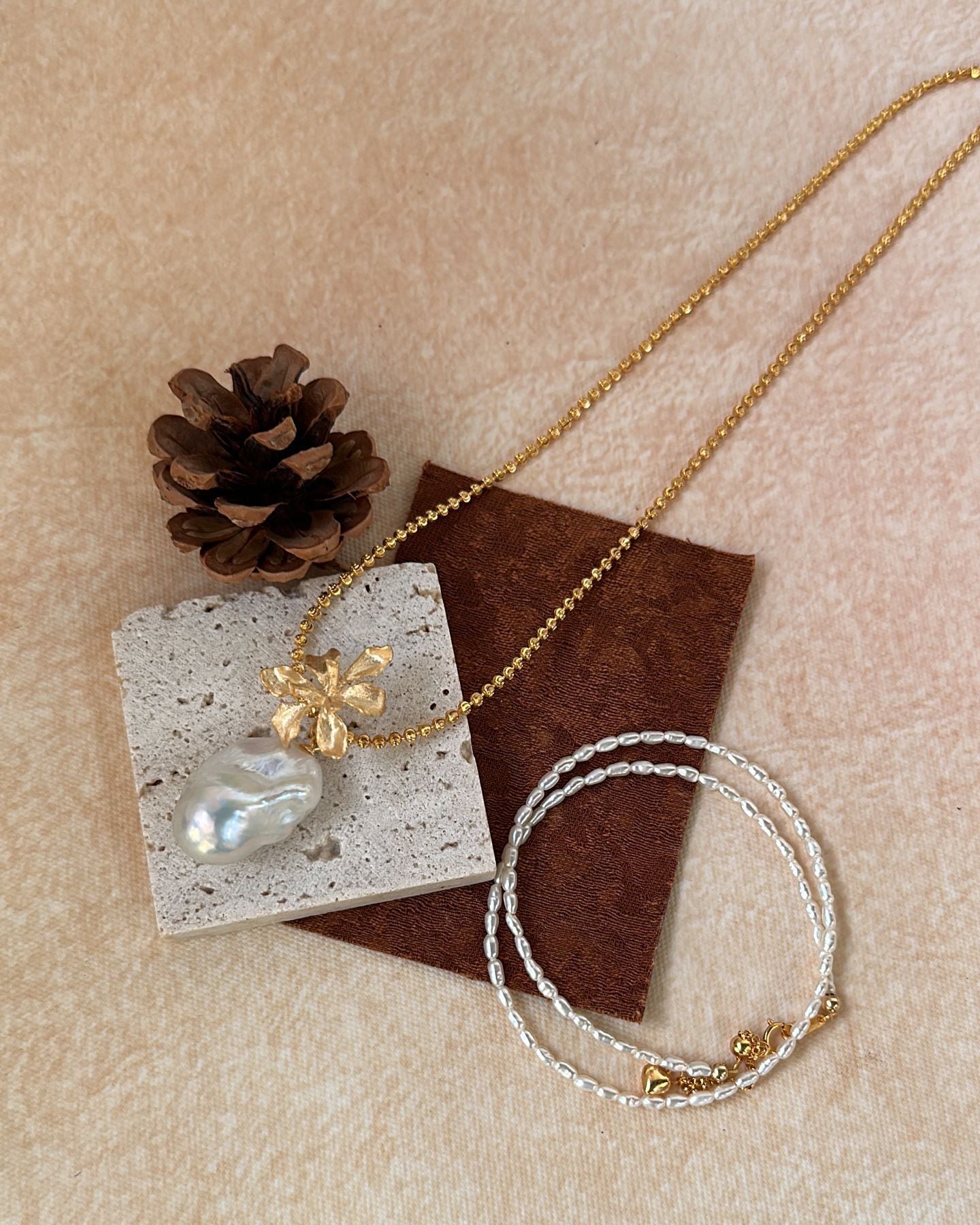 Gold necklace and bracelet on a textured beige surface with a pine cone and card.