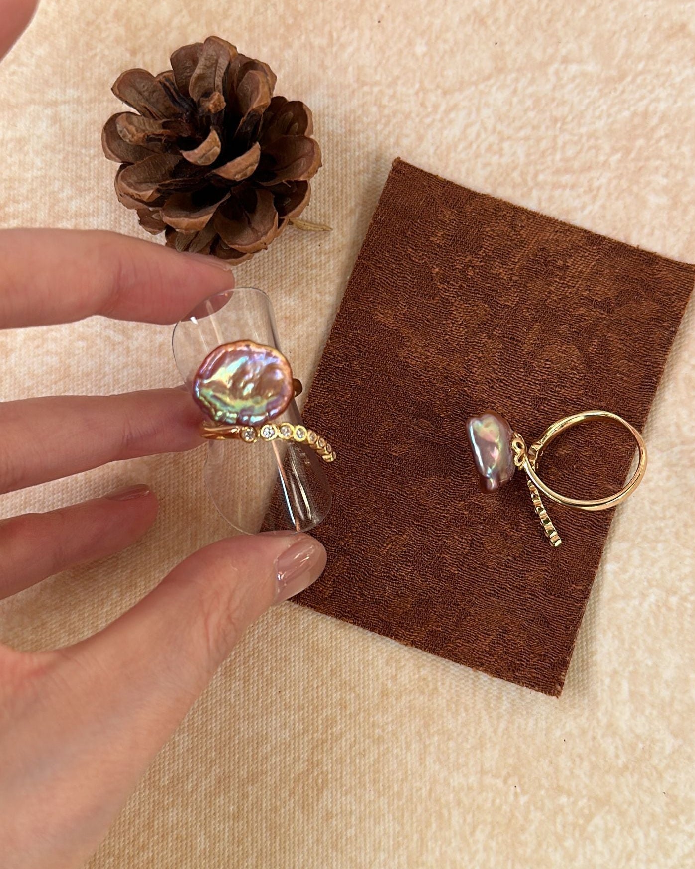 Gold ring with a colorful stone held by a hand, on a brown card with a pine cone and fabric background.