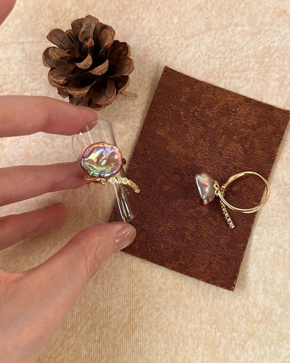 Gold ring with a colorful stone held by a hand, on a brown card with a pine cone and fabric background.