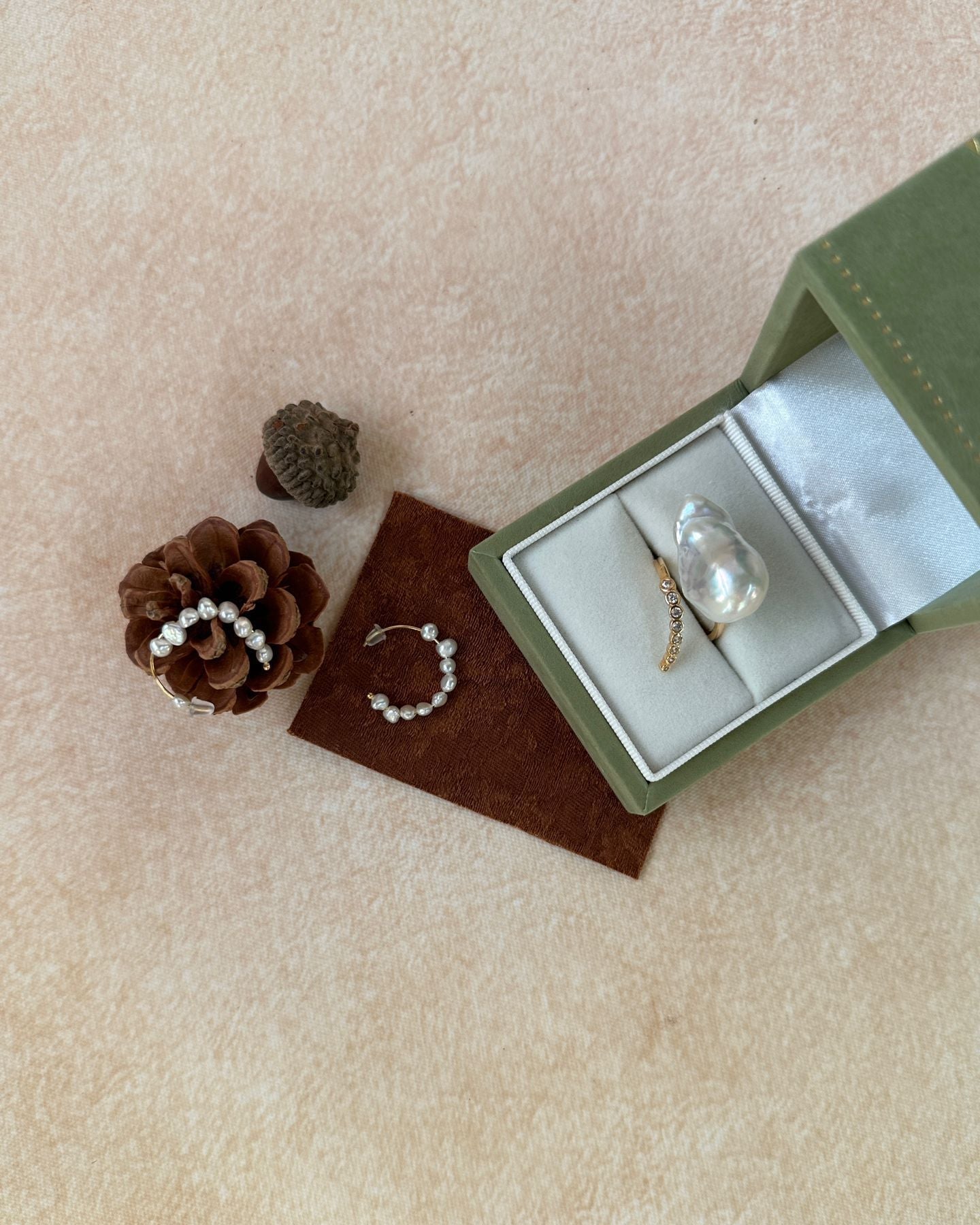 Jewelry set with rings and a bracelet in an open green box on a beige surface with pinecones.