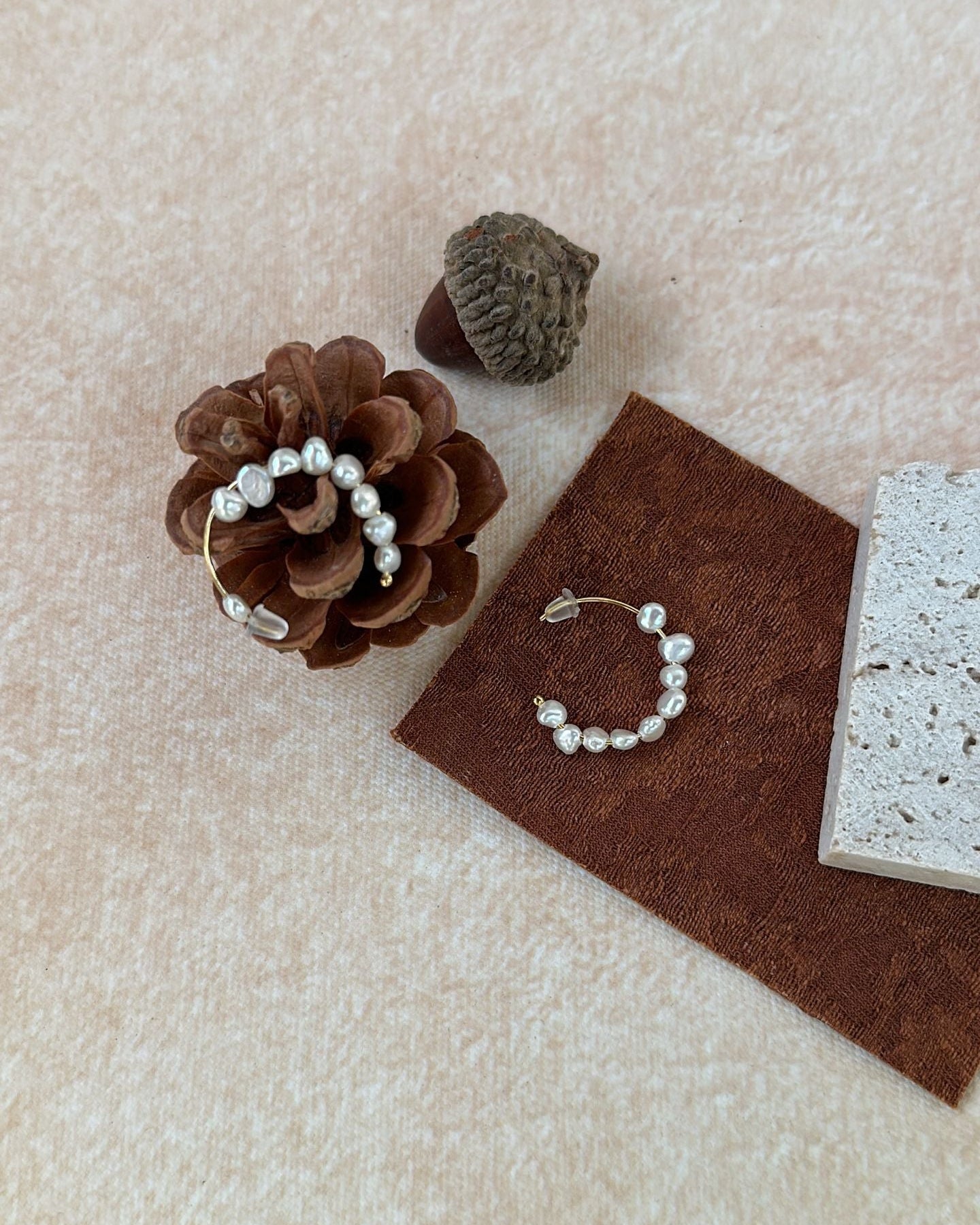 Pearl earring displayed on a pine cone and acorn with a brown fabric square on a beige background
