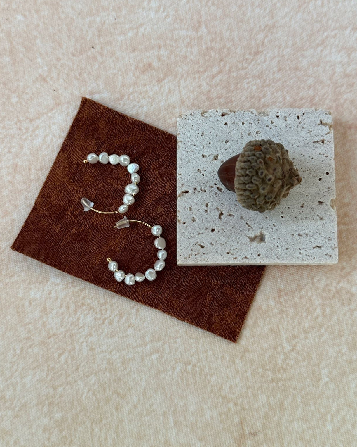 Pearl necklace on a brown fabric square with a stone on a white square, all on a beige surface.