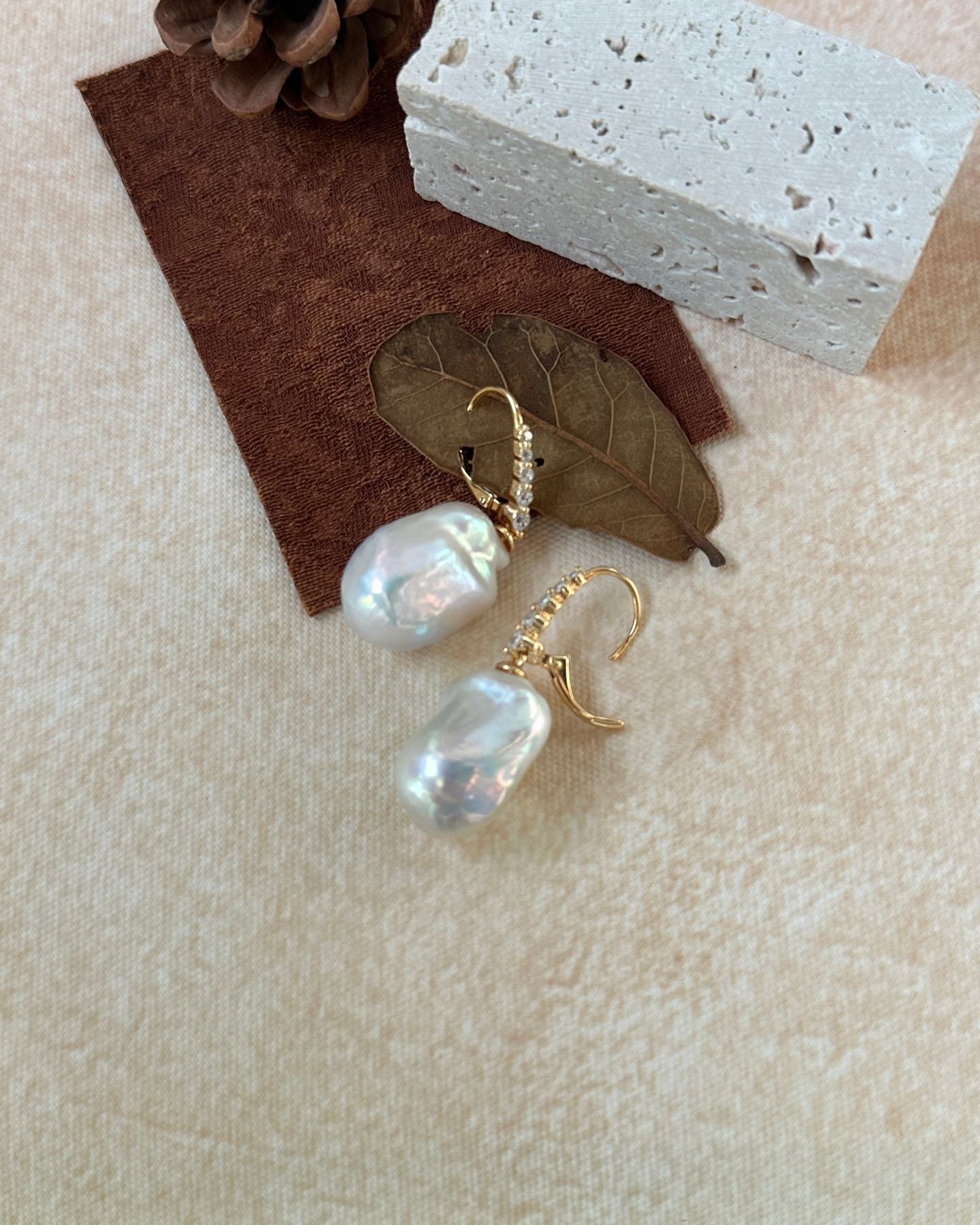 Pearl earrings with gold hooks on a textured surface with a leaf and pine cone.