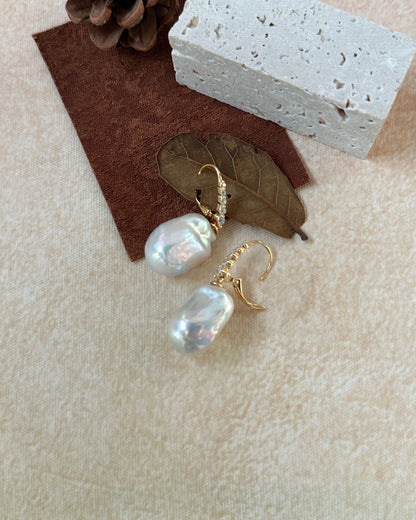 Pearl earrings with gold hooks on a textured surface with a leaf and pine cone.