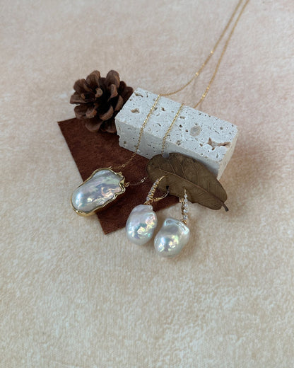 Pearl earrings with a necklace on a textured surface with a pine cone and leaf.