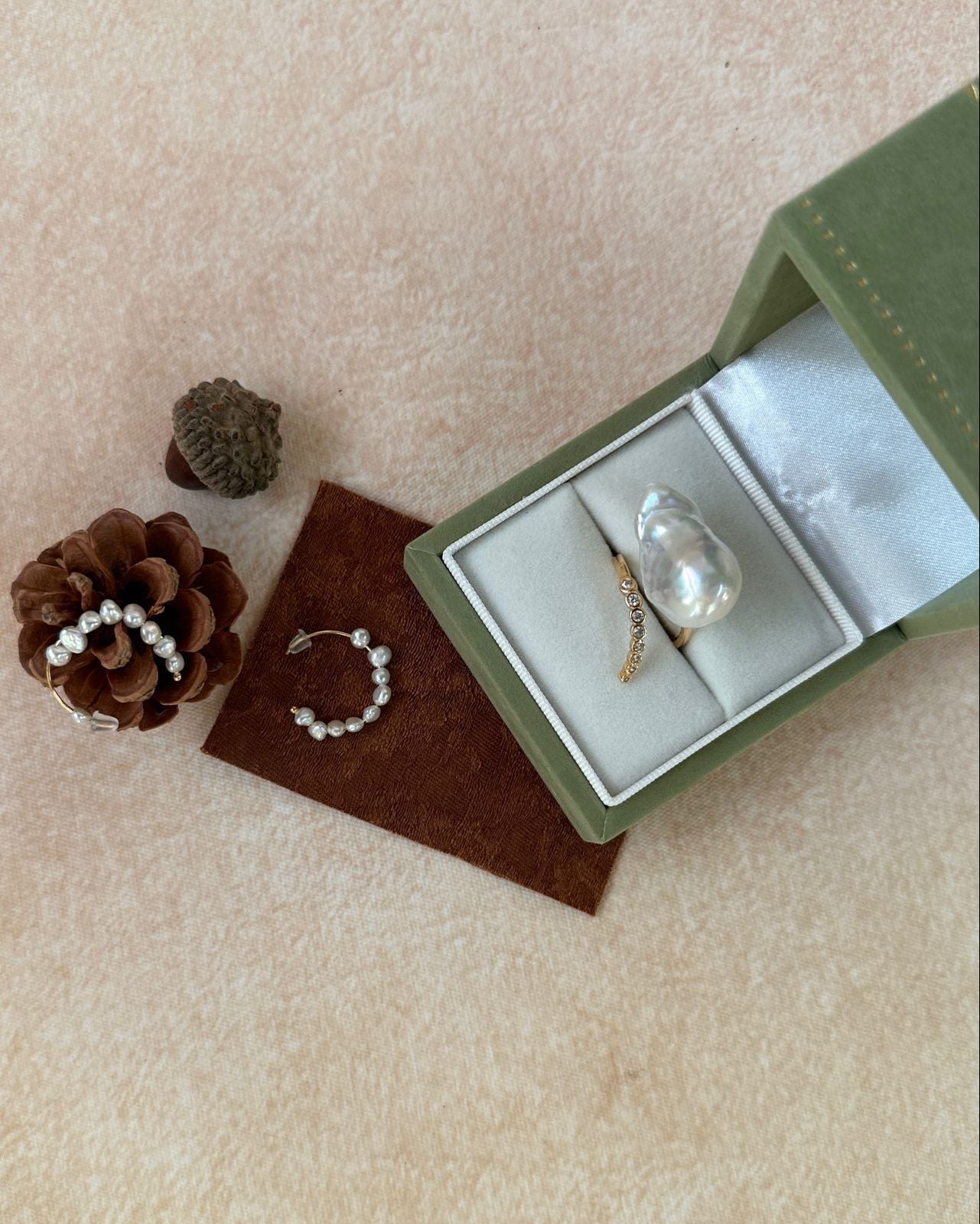 Jewelry set with rings and a bracelet in an open green box on a beige surface with pinecones.