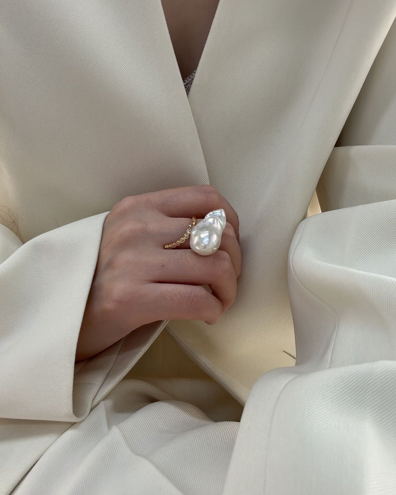 Hand wearing a pearl ring with a white coat in the background