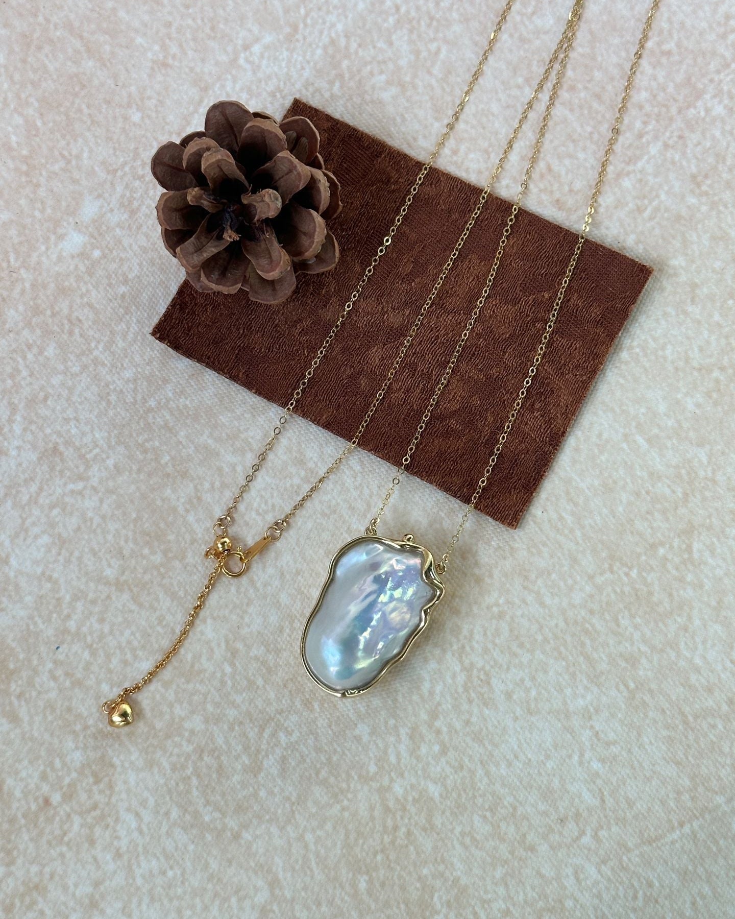 Gold necklace with a large pearl pendant on a brown card with a pine cone, on a beige surface.