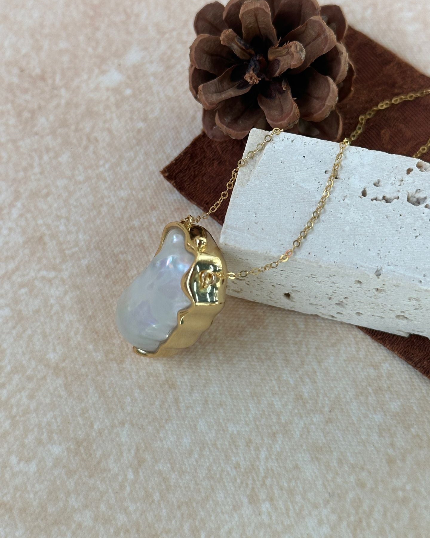 Gold necklace with a pendant on a textured surface with a pine cone in the background