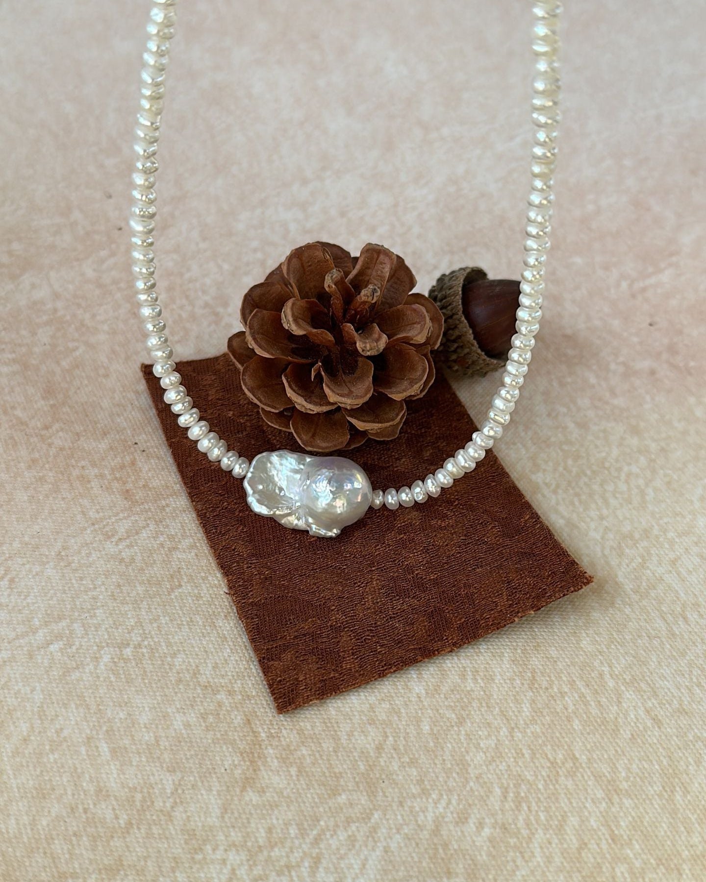 Pearl necklace with a decorative bead on a brown square pad with pinecones.