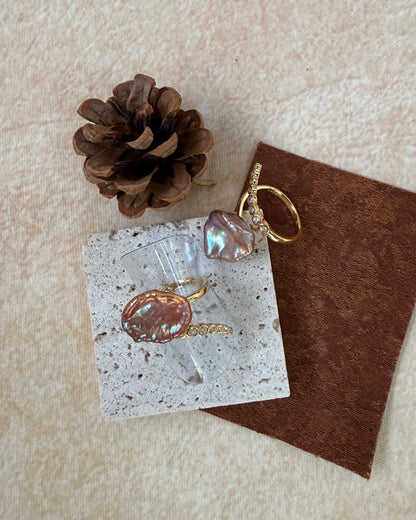 Pair of earrings on a card with a pine cone and brown fabric on a textured beige surface