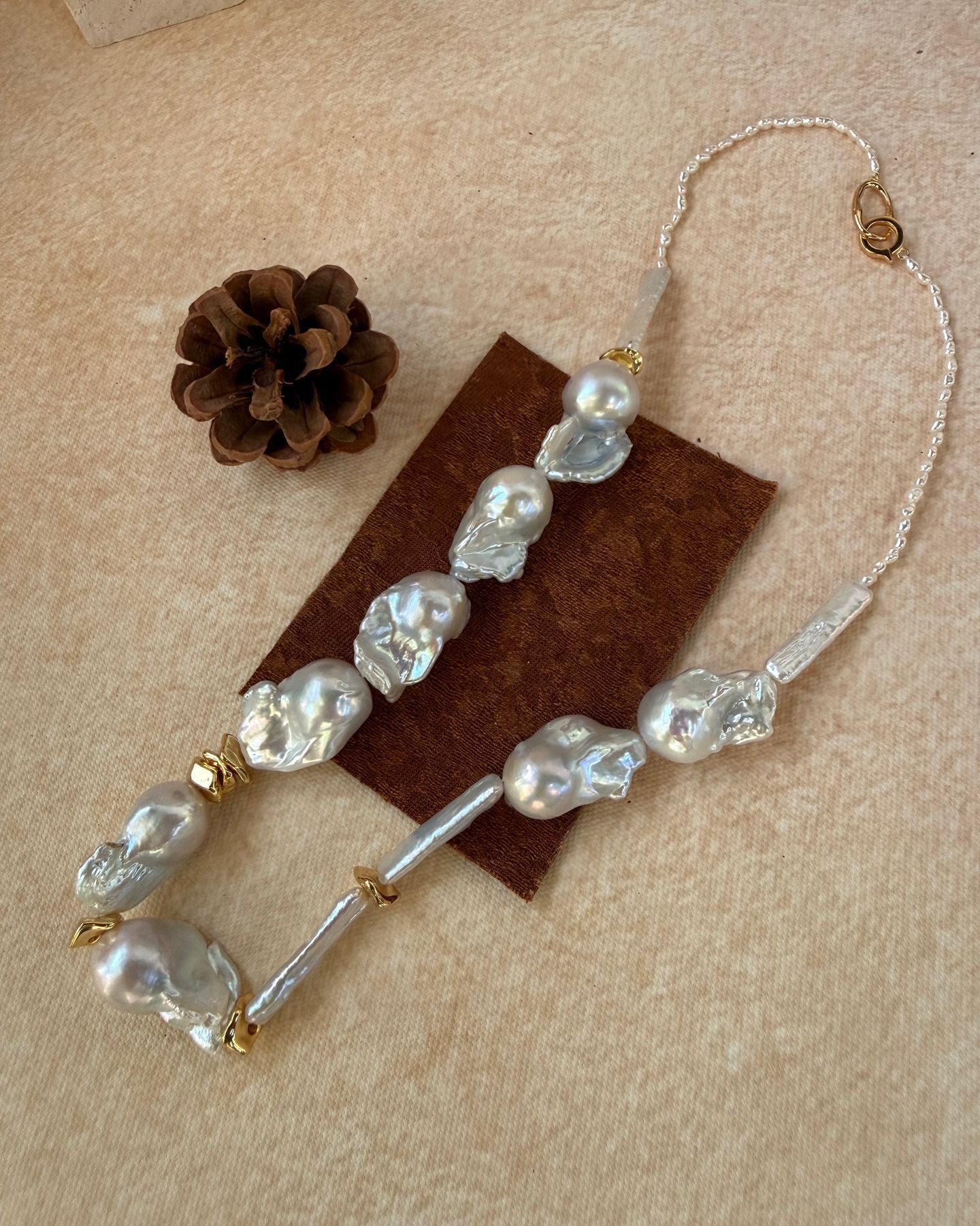 Pearl and gold beaded hair clip on a beige surface with a pine cone and brown fabric.